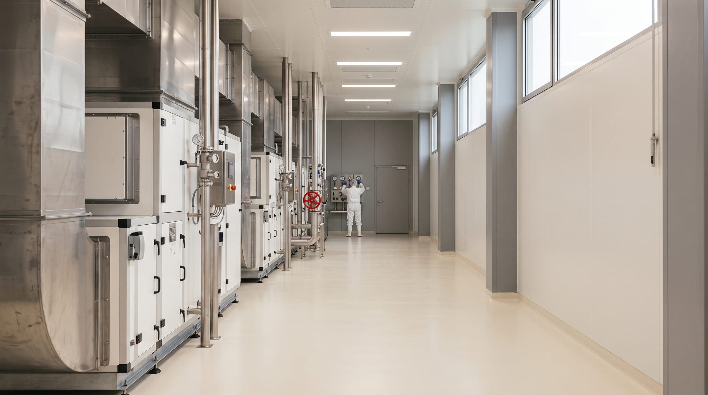 A gowned operator checking gauges at the end of a pharmaceutical process hall lined with air-handling units and stainless ducting.
